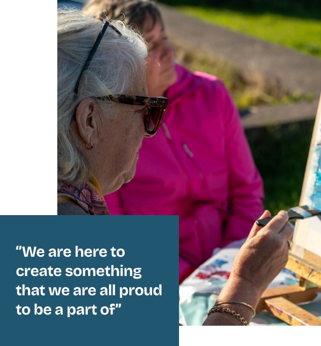 Two elderly women sit outdoors, one wearing sunglasses, painting on canvases. They are focused on their work. A quote overlay reads, "We are here to create something that we are all proud to be a part of" against a blue background.