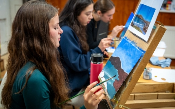 Three women sit in a row, focused on painting landscapes at easels. The first woman, in the foreground, holds a paintbrush and a red thermos. The scene includes blue skies and a mountainous coastline depicted on their canvases.