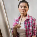 A woman in a pink plaid shirt holds a paintbrush to her chin, looking thoughtful as she sits in front of a blank canvas on an easel. Behind her, a classical bust sculpture is partially visible.