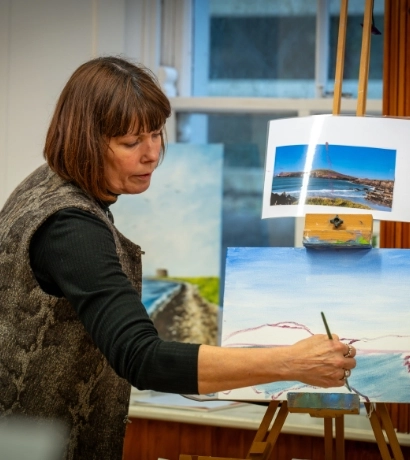 A woman with brown hair is painting on a canvas in a well-lit room. She is wearing a textured vest and a black long-sleeve shirt. A reference photo is clipped to the easel above her artwork.