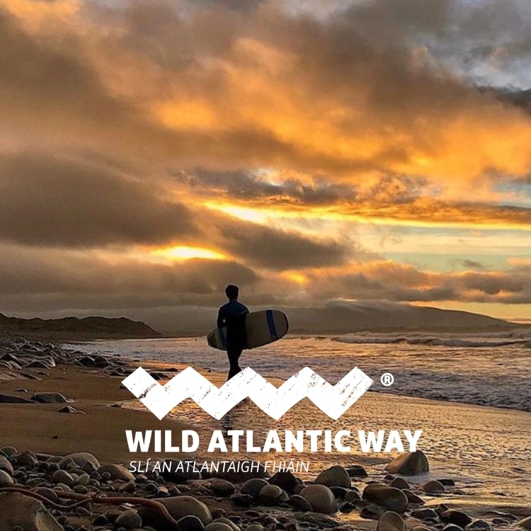 A person holding a surfboard walks towards the sea at sunset, with dramatic clouds and waves in the background. The text reads "Wild Atlantic Way, Sli an Atlantaigh Fhiain." Pebbles and sand cover the shore.